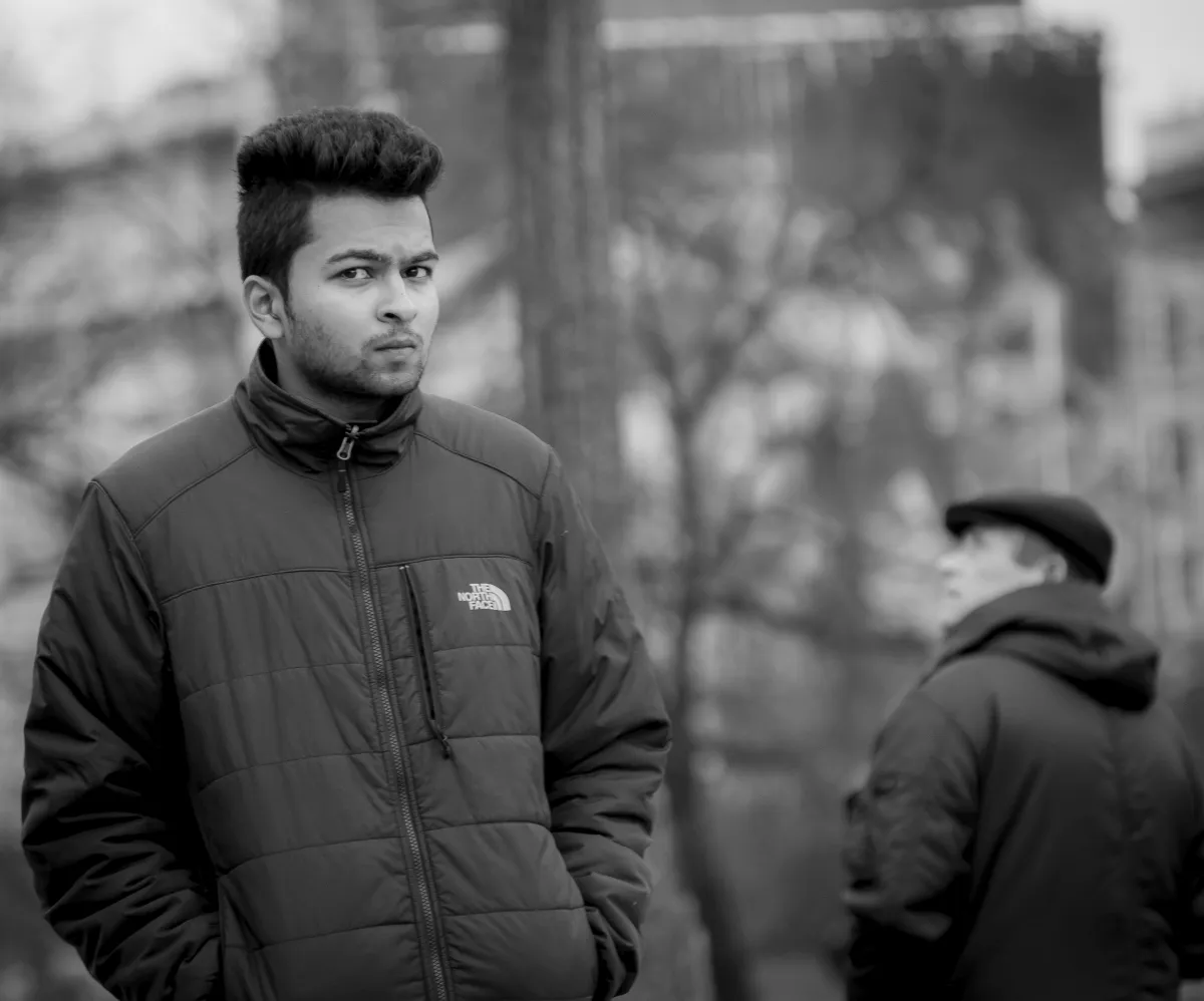 Young man in a quilted jacket staring ahead while an older man stands blurred behind him
